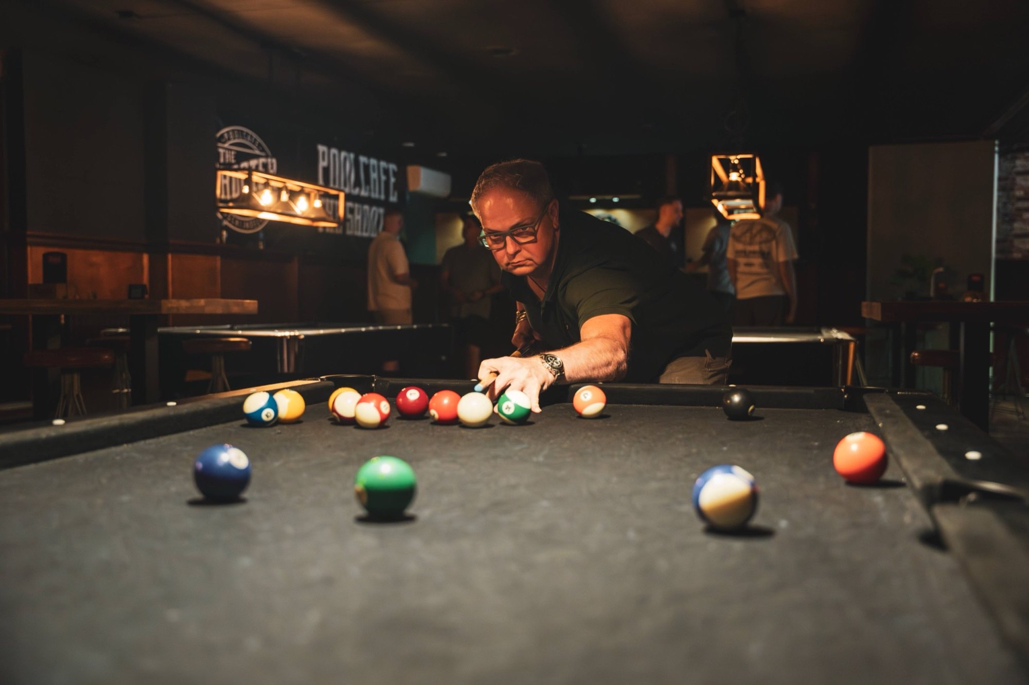Man met bril speelt pool bij The Shooter, met het poolcafe-neonbord op de achtergrond